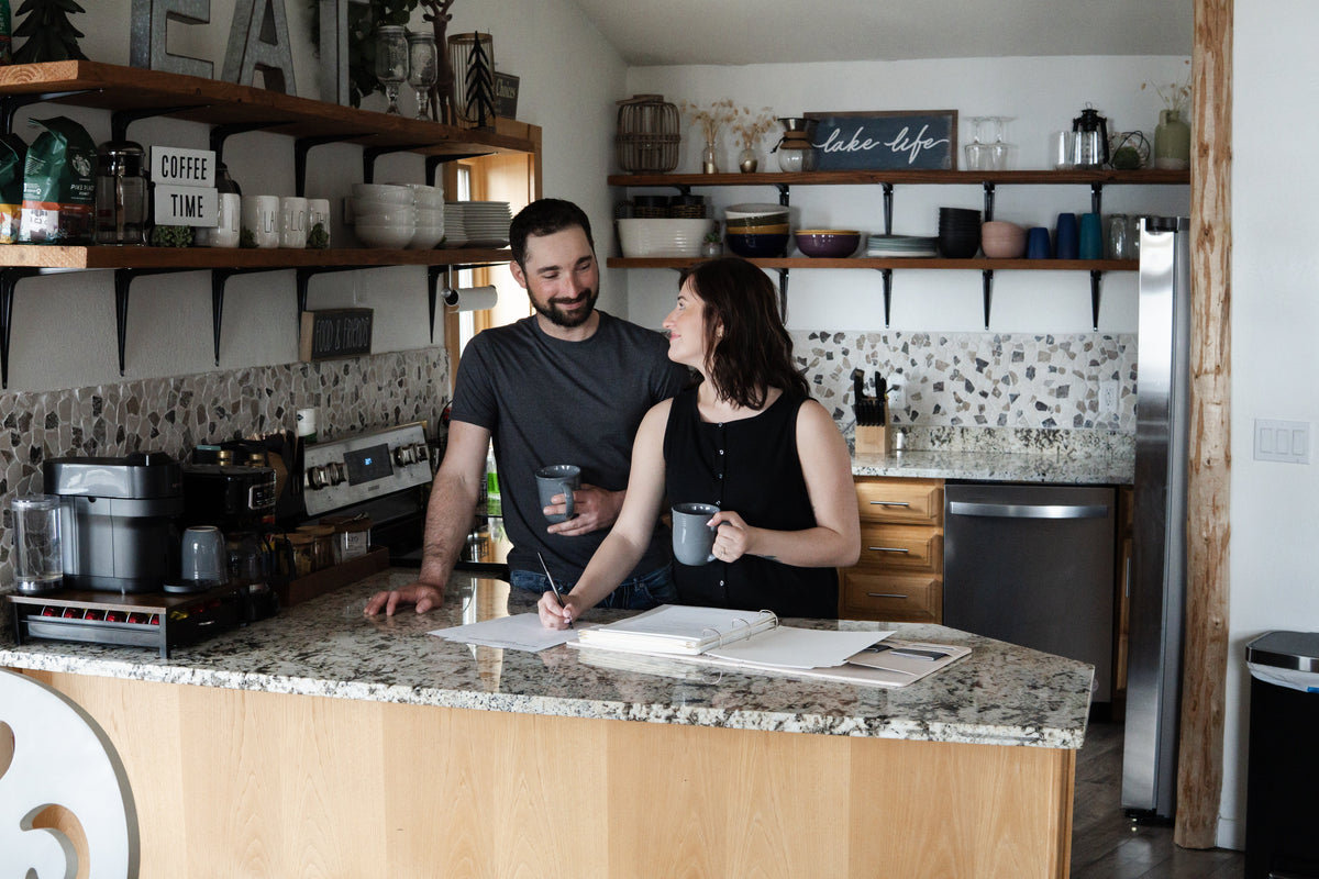 Newlywed couple creating their first monthly budget together at a kitchen table with bills, a laptop, and a notebook.